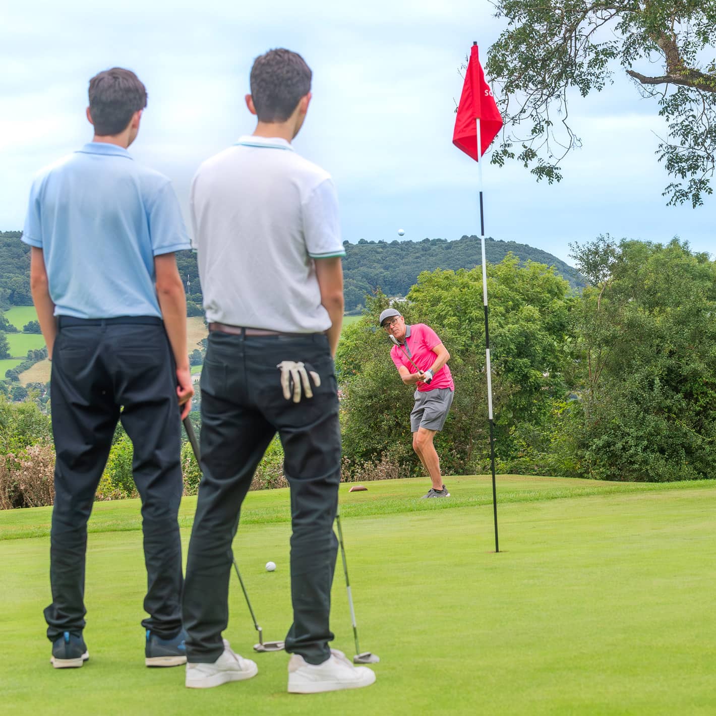 Two golfers watching as another chips a shot towards the hole at Sidmouth Golf Club